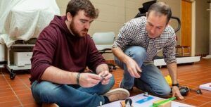 Two men sit on a tiled floor working together on a project using small tools and colorful materials, focusing intently on their task. Chairs and covered equipment are visible in the background.