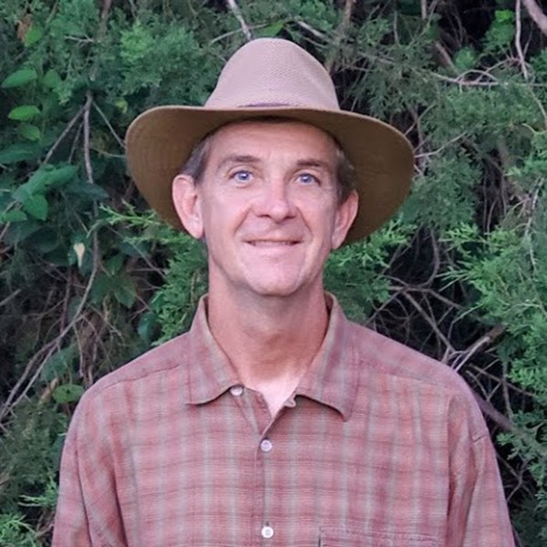 A man wearing a tan wide-brimmed hat and a brown plaid shirt stands outdoors in front of green foliage, smiling at the camera.