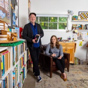 A man stands by a bookshelf holding books, while a woman sits in a leather chair with a tablet in a bright, cozy room filled with books, art supplies, and colorful decorations.