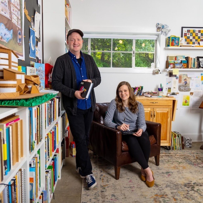 A man stands by a bookshelf holding books, while a woman sits in a leather chair with a tablet in a bright, cozy room filled with books, art supplies, and colorful decorations.