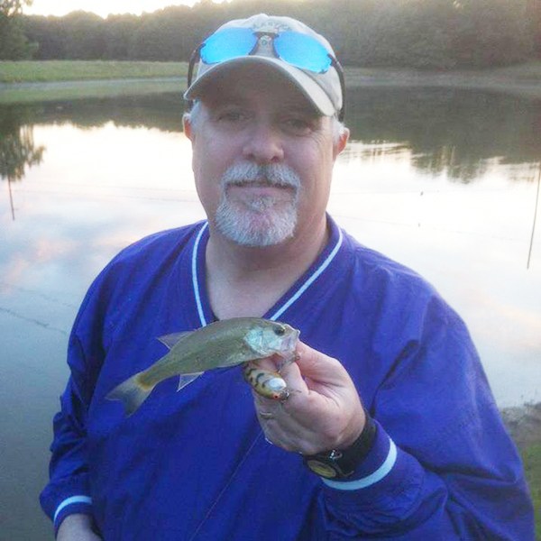 A man in a blue jacket and beige cap stands by a pond, smiling and holding a small fish in his hand. The pond and trees are visible in the background during daylight.