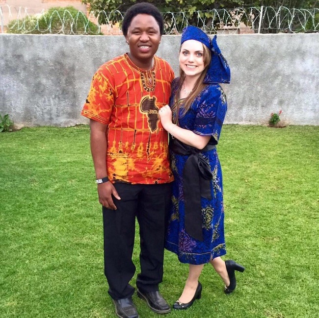 A smiling man and woman stand together on grass, wearing colorful traditional African attire. The man wears an orange patterned shirt and black pants; the woman wears a blue dress with a matching headwrap. A stone wall is behind them.