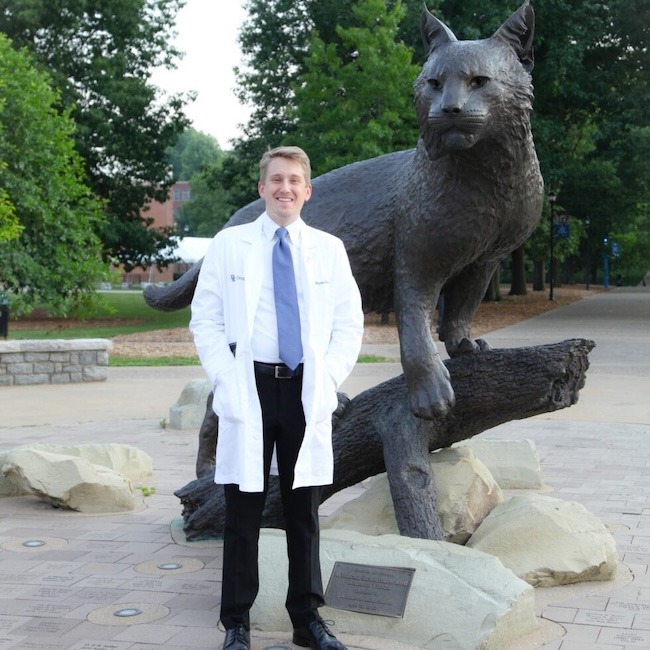 A man in a white lab coat stands smiling in front of a large wildcat statue outdoors, surrounded by trees and stone plaques.
