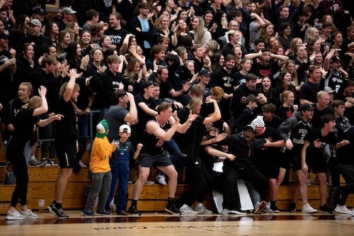 A large, enthusiastic crowd of students wearing mostly black cheers and celebrates in the bleachers at an indoor sports event. One child in a costume stands out among the crowd.