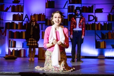A young woman kneels on stage, smiling and holding a white scarf, with two people standing behind her against a backdrop of bookshelves and dramatic lighting, suggesting a theatrical performance.
