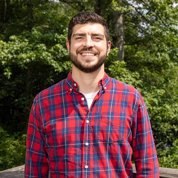 A man with short dark hair and a beard, wearing a red and blue plaid shirt, stands outdoors in front of green trees, smiling at the camera.