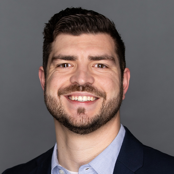 Smiling man with short dark hair and a trimmed beard, wearing a navy blazer and light blue collared shirt, posing in front of a plain gray background.