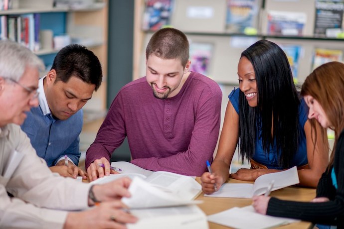 Five adults sit together at a table in a library or classroom, reading books and taking notes, appearing engaged and smiling as they collaborate on a group study session. Shelves with magazines are visible in the background.