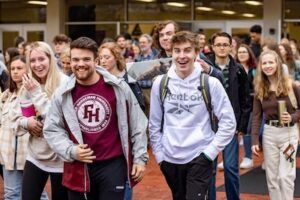A group of smiling young adults, some holding books and backpacks, walk together outdoors in front of a building with glass doors. The atmosphere appears friendly and energetic.