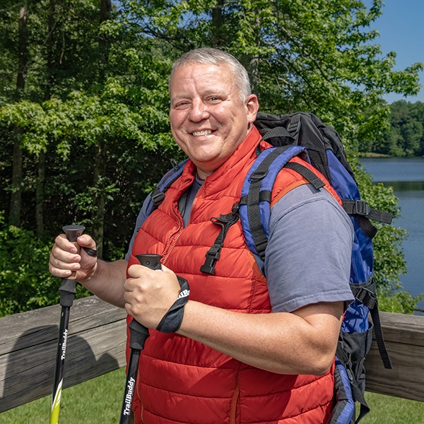 A smiling person with short gray hair wearing a red vest and backpack holds trekking poles on a wooden bridge, with trees and a lake in the background on a sunny day.