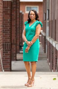 A woman in a green dress and light pale orange heels stands confidently outdoors between brick buildings, smiling with her hands clasped in front of her.