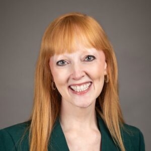 A smiling woman with straight red hair, wearing hoop earrings and a blue top, poses in front of a plain gray background.