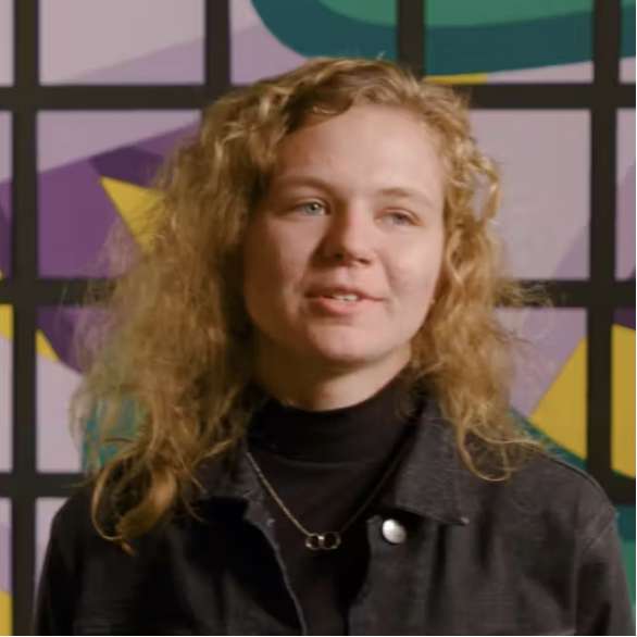 A person with curly blonde hair, wearing a black turtleneck and dark jacket, stands in front of a colorful geometric patterned background.