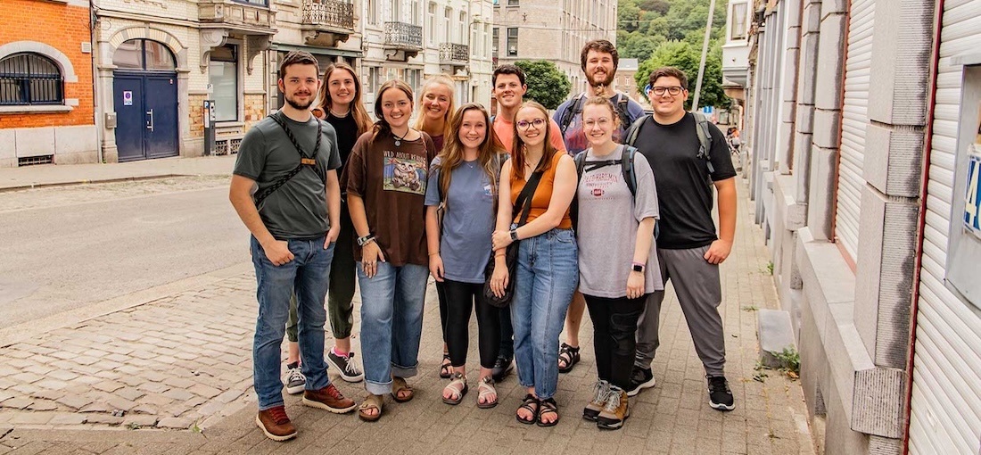 A group of ten young adults stand closely together, smiling at the camera on a city sidewalk with old buildings and greenery in the background.