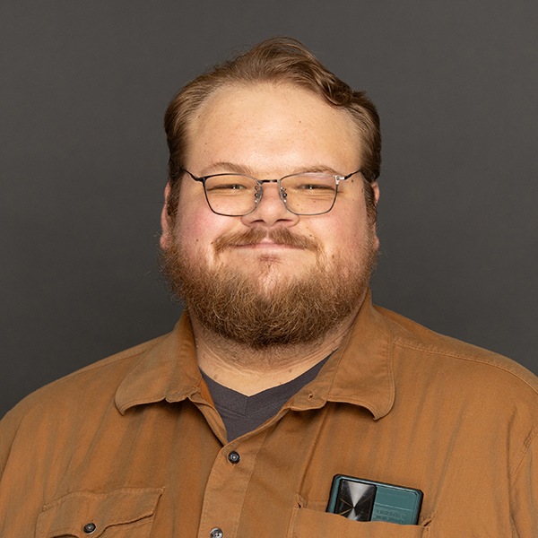 A smiling man with glasses, a beard, and light brown hair wearing a brown button-up shirt with a pen and a pocket protector in the chest pocket, against a plain dark background.