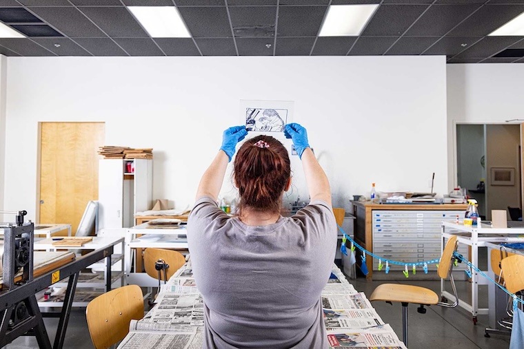 A person with brown hair and blue gloves examines a photo or print, holding it up in a bright, organized art studio with long tables covered in newspapers. Their back faces the camera.