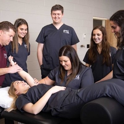 Six people in medical scrubs stand around a woman lying on a treatment table. One person assists her arm while others observe, suggesting a physical therapy or medical training session. Everyone appears focused and engaged.