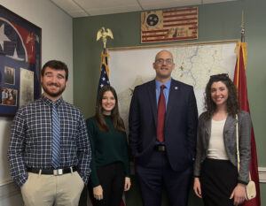 Four people stand smiling in an office with maps and American and Tennessee flags on the wall behind them. Three are casually dressed and one man in the center wears a suit and tie.