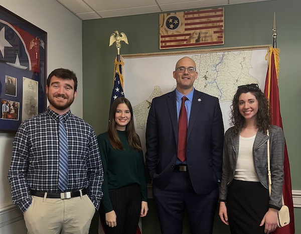 Four people stand smiling in an office with maps and American and Tennessee flags on the wall behind them. Three are casually dressed and one man in the center wears a suit and tie.