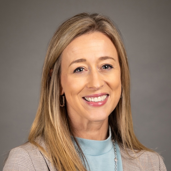 Smiling woman with shoulder-length blonde hair, wearing a black blazer over a patterned top, and small earrings, posing in front of a plain gray background.
