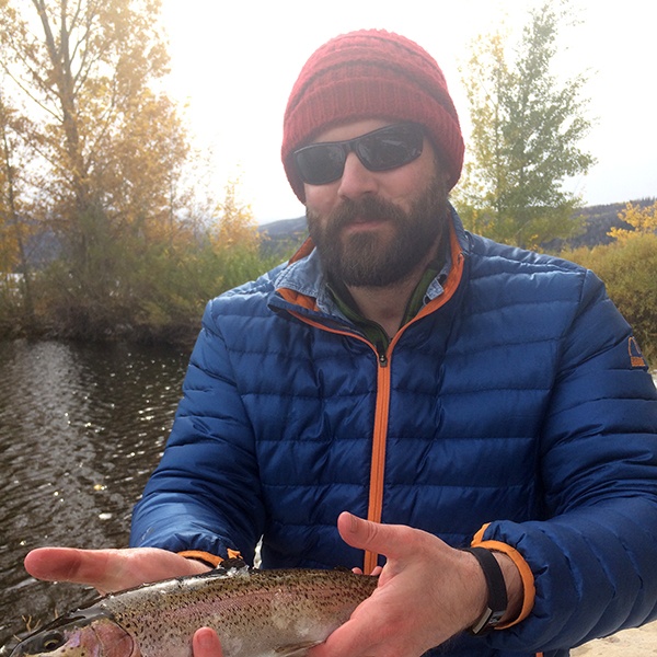 A bearded man in a blue jacket, red beanie, and sunglasses holds a fish with both hands by a lake, with autumn trees and water in the background.