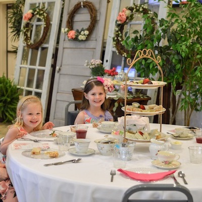 Two young girls in floral dresses sit smiling at a round table set for tea, with tiered trays of treats and teacups. The background features vintage doors decorated with wreaths and flowers.