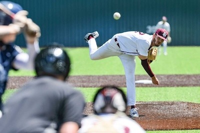 A baseball pitcher in a white uniform throws a pitch toward the batter, while the catcher and umpire watch from behind home plate on a sunny field.