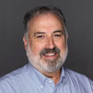 A middle-aged man with gray hair and a beard smiles while wearing a light blue, striped button-down shirt against a plain dark gray background.