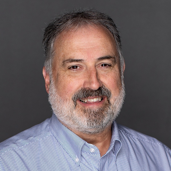 A middle-aged man with gray hair and a beard smiles while wearing a light blue, striped button-down shirt against a plain dark gray background.