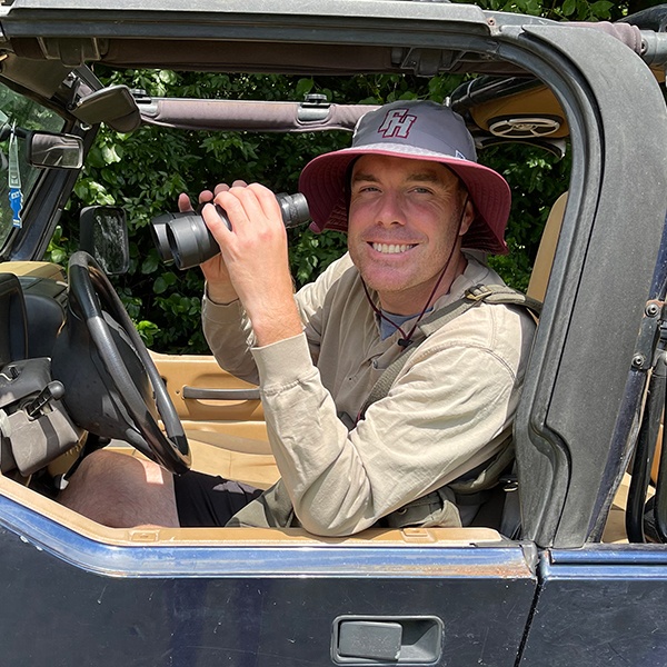 A smiling man wearing a wide-brimmed hat and beige shirt sits in a blue open-top vehicle, holding binoculars, with green foliage in the background.