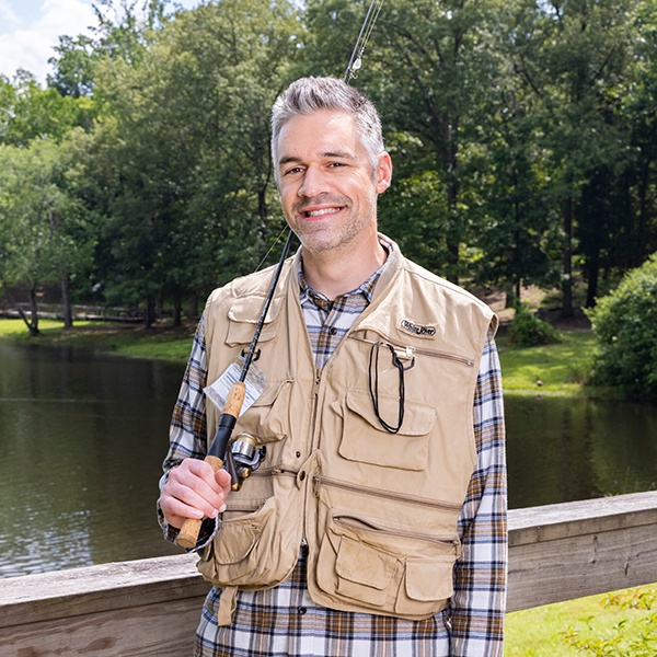 A smiling man in a plaid shirt and beige fishing vest holds a fishing rod and stands on a bridge by a pond, with trees and greenery in the background.