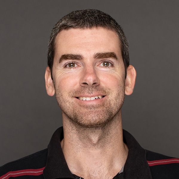 A man with short brown hair and trimmed beard, wearing a black collared shirt with red stripes, smiles at the camera against a plain gray background.