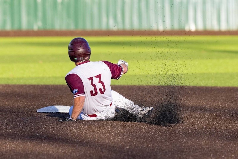 A baseball player wearing a maroon helmet and jersey number 33 slides into a base on a dirt infield, kicking up a spray of dirt, with a green outfield and fence in the background.