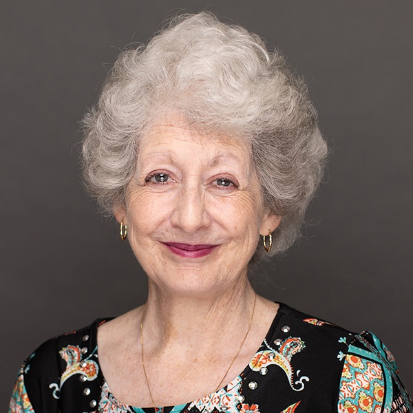 Elderly woman with short, curly gray hair, smiling gently. She is wearing gold hoop earrings, a gold necklace, and a colorful patterned blouse. The background is plain and dark gray.