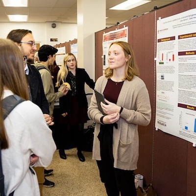 A woman presents a scientific poster to a group of people in a conference setting. She holds a tablet and engages with attendees, while other posters and participants are visible in the background.