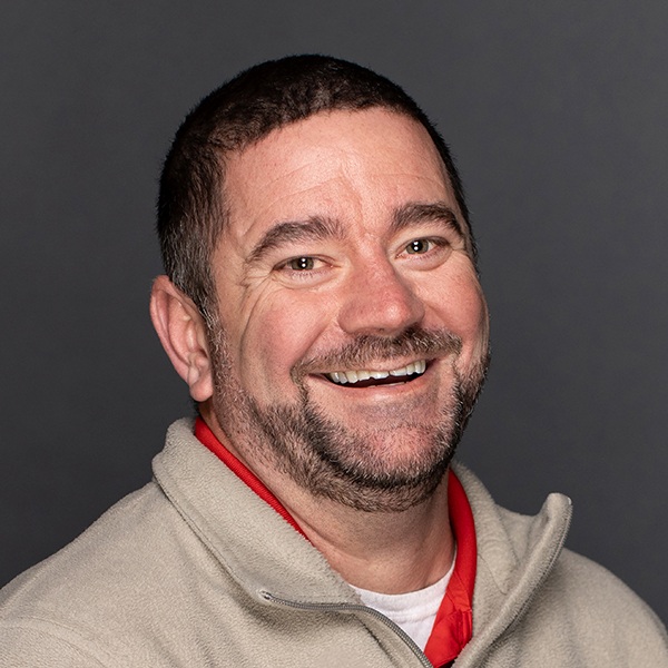 A man with short dark hair and a beard smiles broadly, wearing a beige fleece jacket over a red shirt, against a plain dark background.