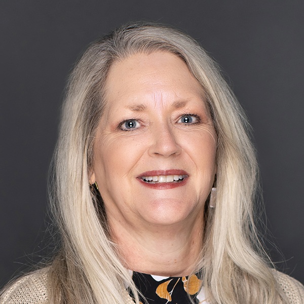 A middle-aged woman with long, straight, light blonde hair smiling at the camera. She is wearing a black top with a patterned collar, a light cardigan, and has a neutral dark background.