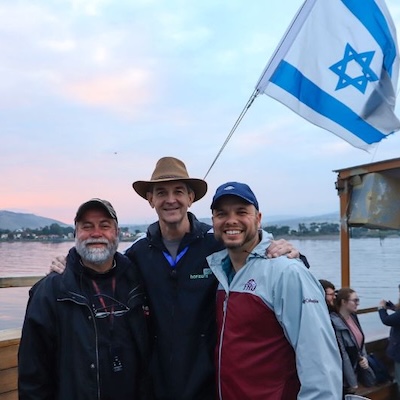 Three smiling men pose in from of a Israel flag on a boat.
