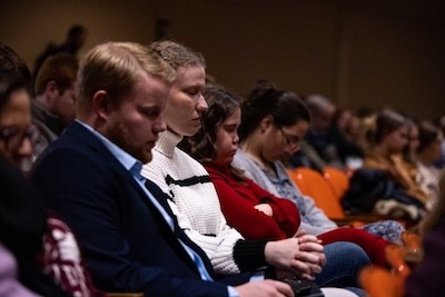 A group of people sit closely together in rows of auditorium seats with their heads bowed and eyes closed, appearing to be in prayer or quiet reflection.