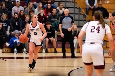 A basketball player in a white LIONS uniform dribbles the ball while a teammate and an opponent face her on the court; spectators and a referee are visible in the background.