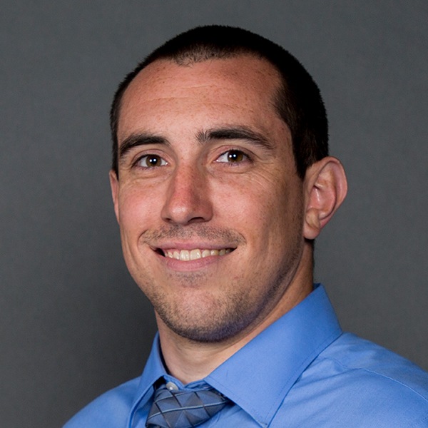 A man with short dark hair and a trimmed beard is smiling at the camera. He is wearing a light blue dress shirt and a patterned gray tie, against a plain dark background.