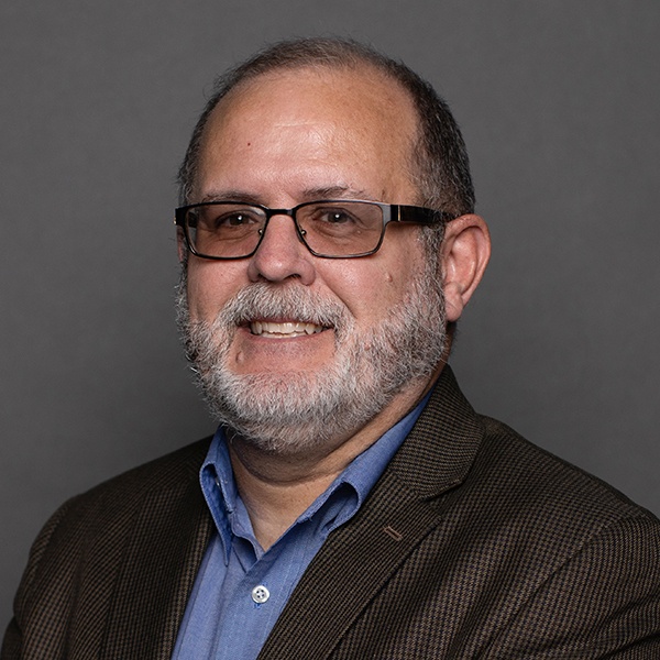 A middle-aged man with glasses, short hair, and a gray beard, wearing a dark plaid blazer over a blue dress shirt, smiles in front of a plain gray background.