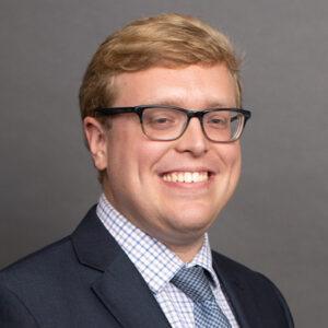 Smiling man with short blond hair and glasses, wearing a dark suit jacket, checked dress shirt, and patterned tie, posing in front of a plain gray background.