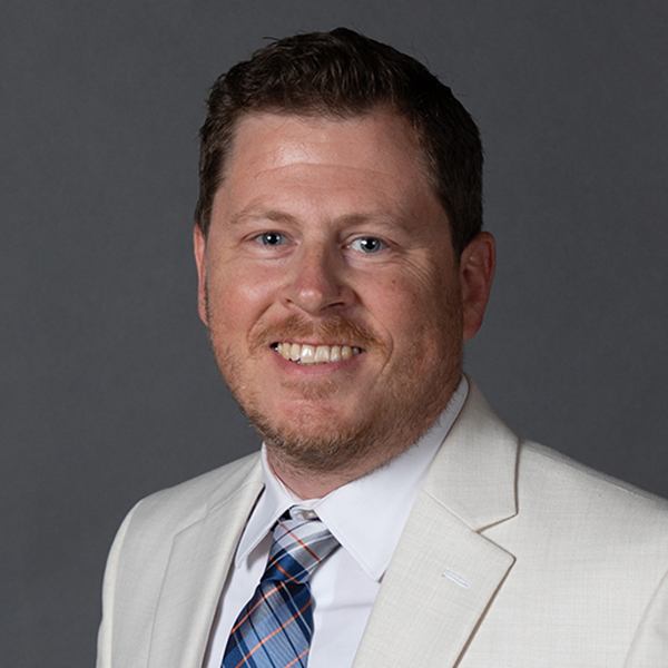 A man with short brown hair and a trimmed beard smiles while wearing a white suit jacket, white dress shirt, and a blue-and-red plaid tie against a plain gray background.