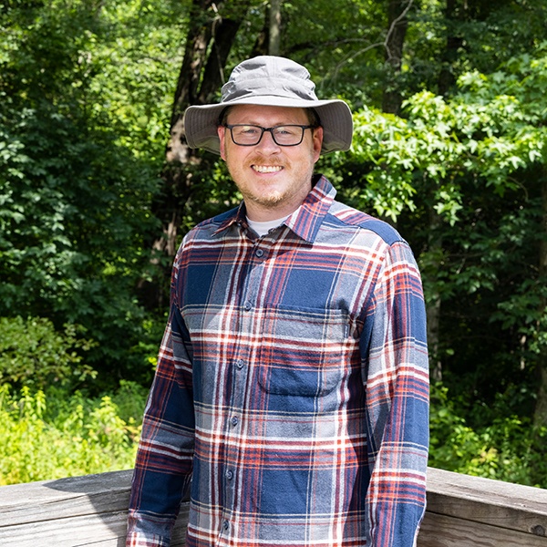 A man wearing glasses, a gray sun hat, and a blue and red plaid shirt smiles while standing on a wooden deck in front of trees and greenery on a sunny day.