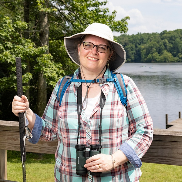 A smiling woman wearing glasses, a sun hat, backpack, and plaid shirt stands by a lake holding a walking stick and binoculars, with trees and water in the background.