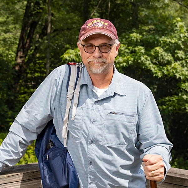 An older man with glasses and a gray beard, wearing a maroon cap, light blue button-up shirt, and backpack, stands outdoors by a wooden railing with green trees in the background, holding a walking stick.