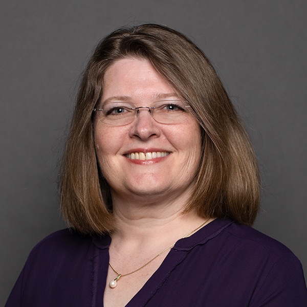 A woman with shoulder-length brown hair, glasses, and a purple blouse, smiling at the camera against a plain gray background.