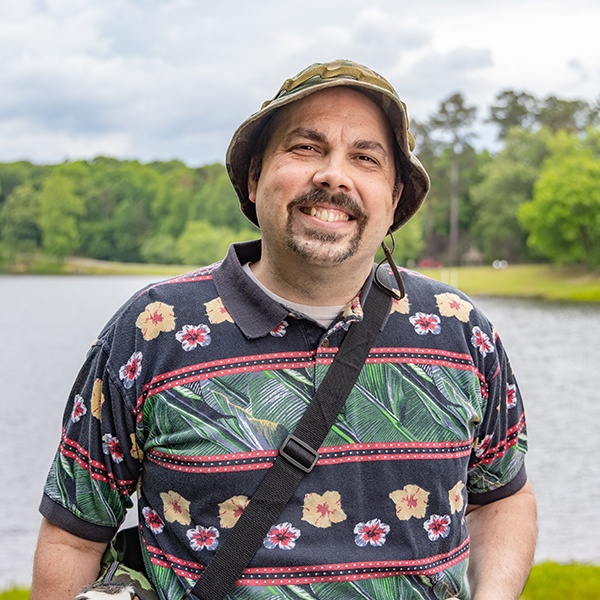 A smiling man with a mustache and beard stands by a lake, wearing a floral-patterned shirt, a camo bucket hat, and a shoulder bag. Green trees and cloudy skies are visible in the background.