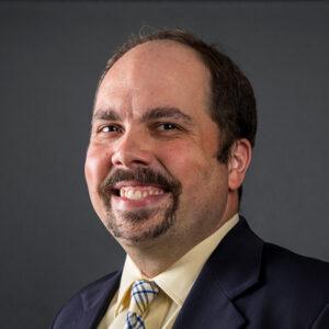 A man with short brown hair, a mustache, and a goatee, wearing a dark suit, light yellow shirt, and striped tie, smiles at the camera against a plain dark background.